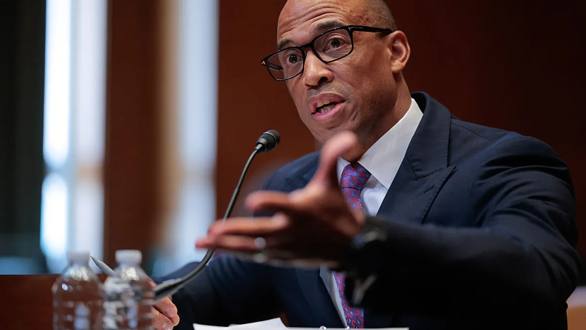 A seated person wearing a suit and glasses gestures while speaking into a microphone.