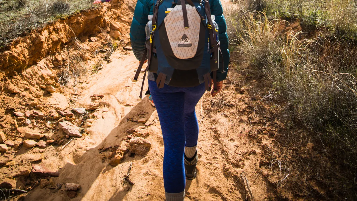 Two people hiking single file toward massive rock formations, seen from the back, with a backpack on the person behind.