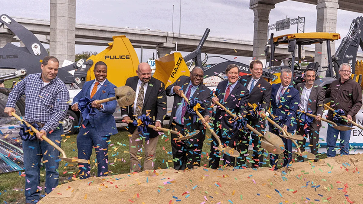 A row of nine men, mostly wearing suits, stand with shovels in front of a pile of dirt. There is construction equipment in the background and confetti raining down.