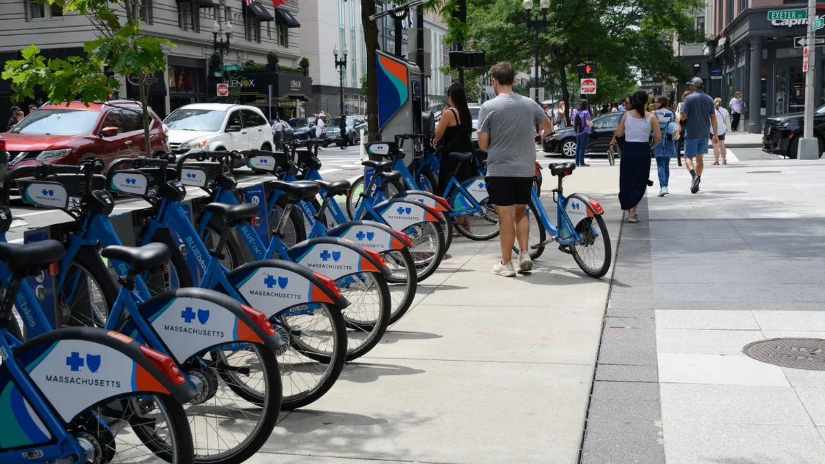 People with a row of docked bicycles along a busy sidewalk.