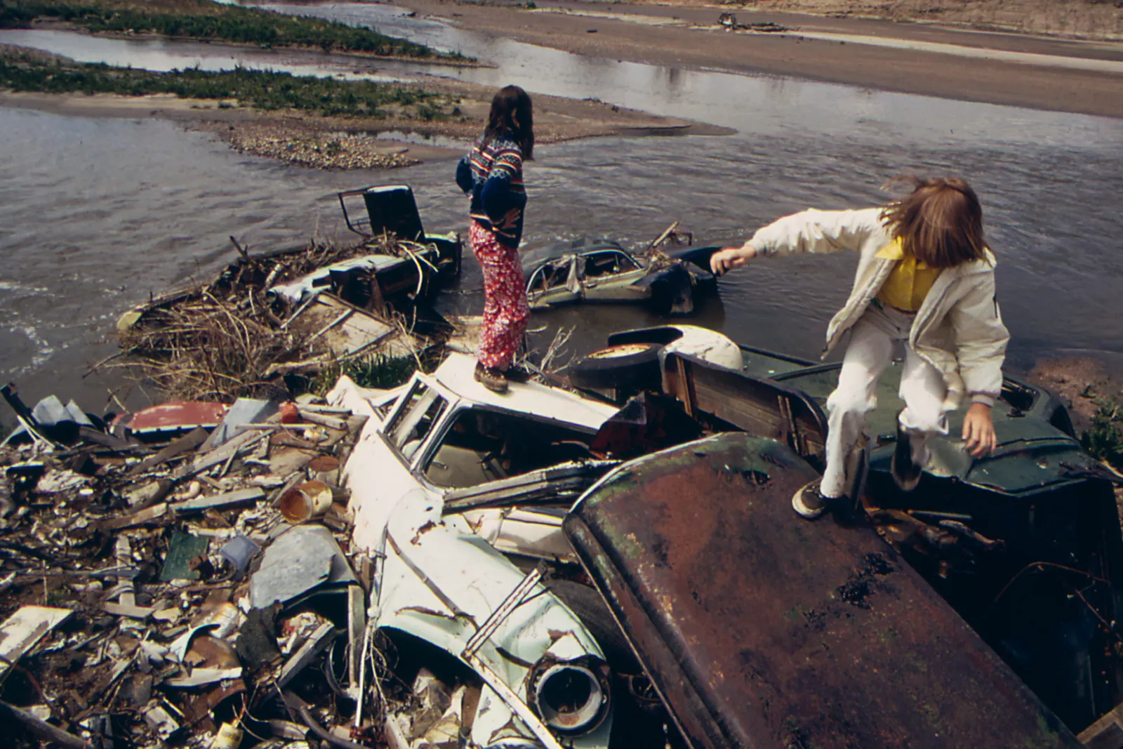 Two children stand on abandoned cars sinking into a body of water