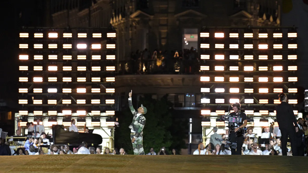 Pharrell Williams stands at the back of a runway and acknowledges the applause of the audience while wearing a camoflage suit.