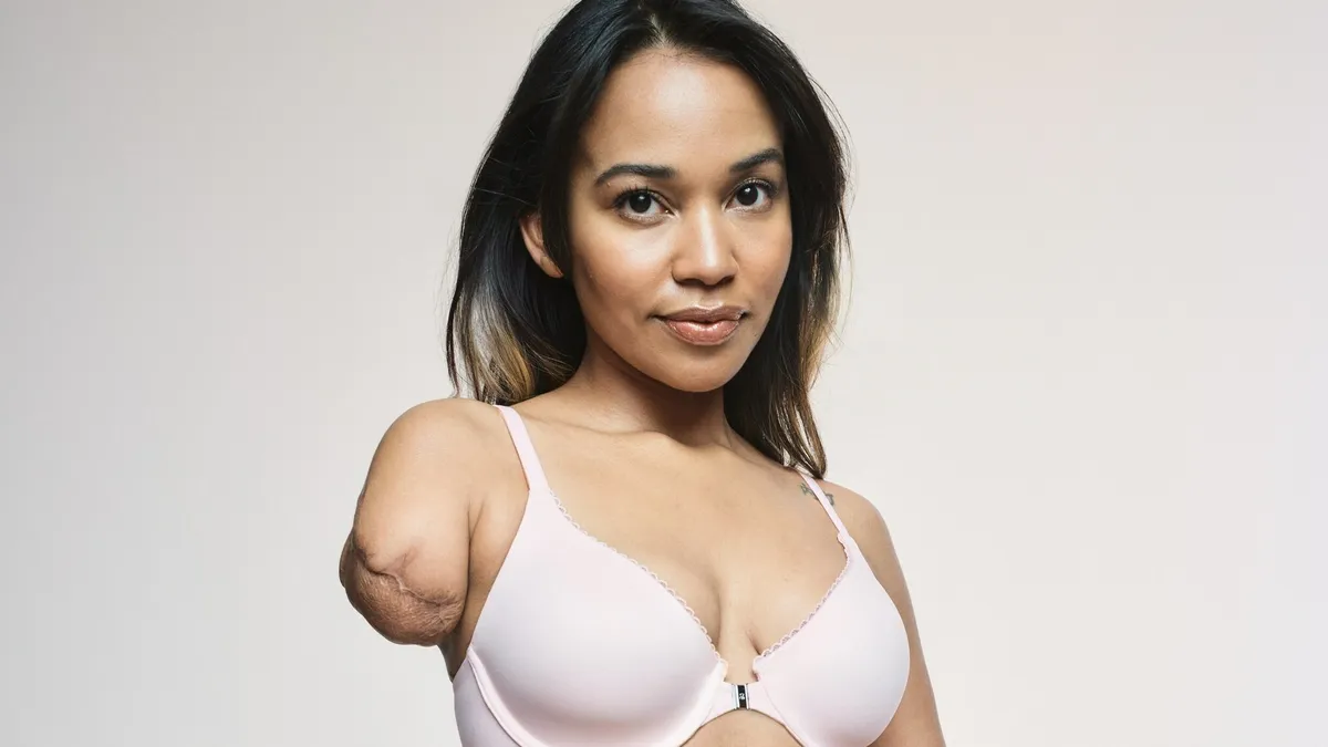 A woman stands and wears a cream-colored bra and underwear from the Victoria's Secret line of adaptive clothing. She is a left-arm amputee.