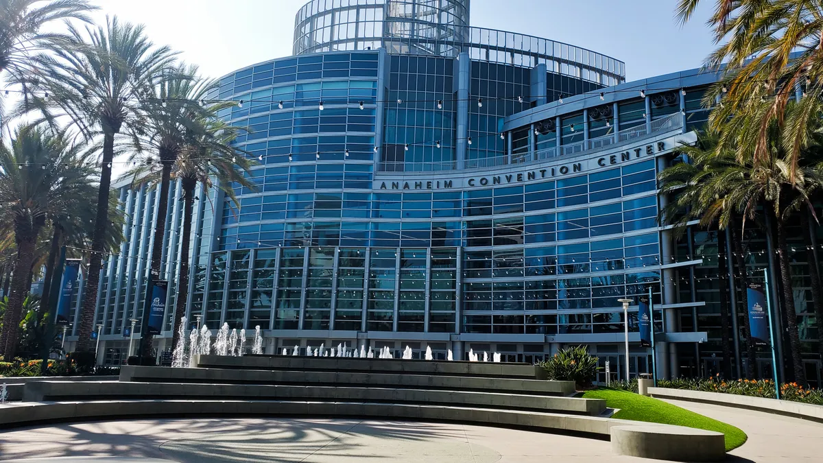 A glass building that says "Anaheim Convention Center' is surrounded by palm trees with a fountain in the front.