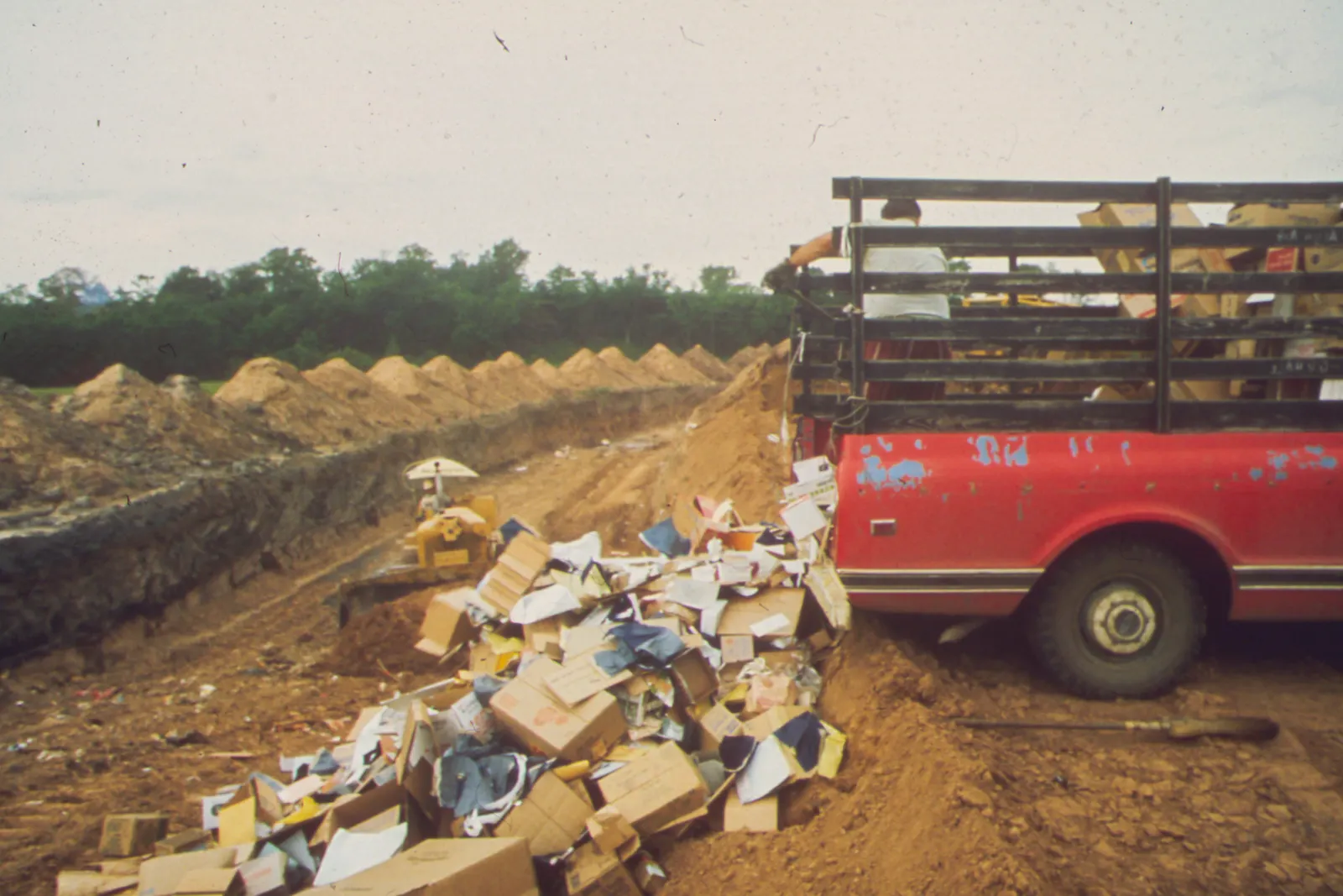 A person stands in the back of a pickup truck, shoveling waste into a landfill