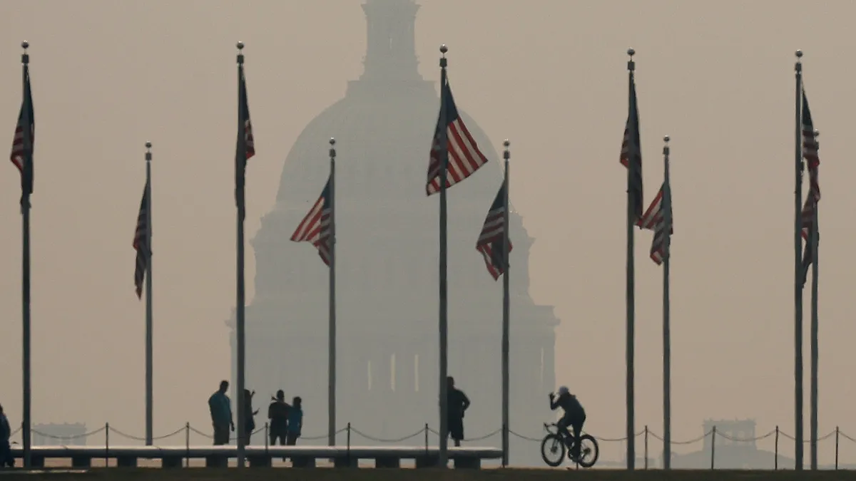 Several pedestrians and a cyclist are silhouetted against a gray sky. A large building looms in the background. Flag poles with the U.S. flag sit in the foreground.