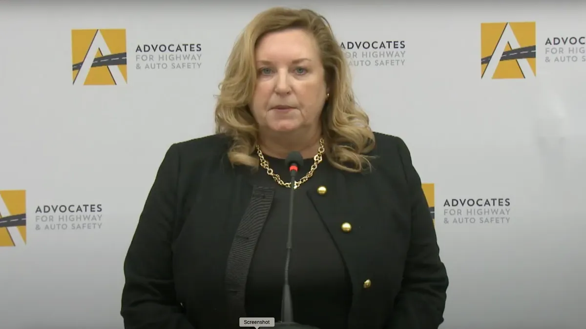 A woman dressed in black standing at a podium with signage behind reading "Advocates for Highway & Auto Safety"