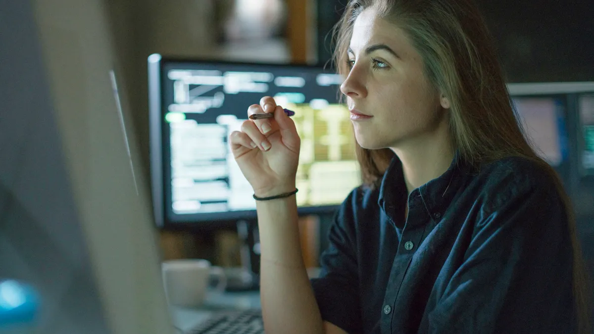 A young woman is seated at a desk surrounded by monitors displaying data, she is contemplating in this dark, moody office