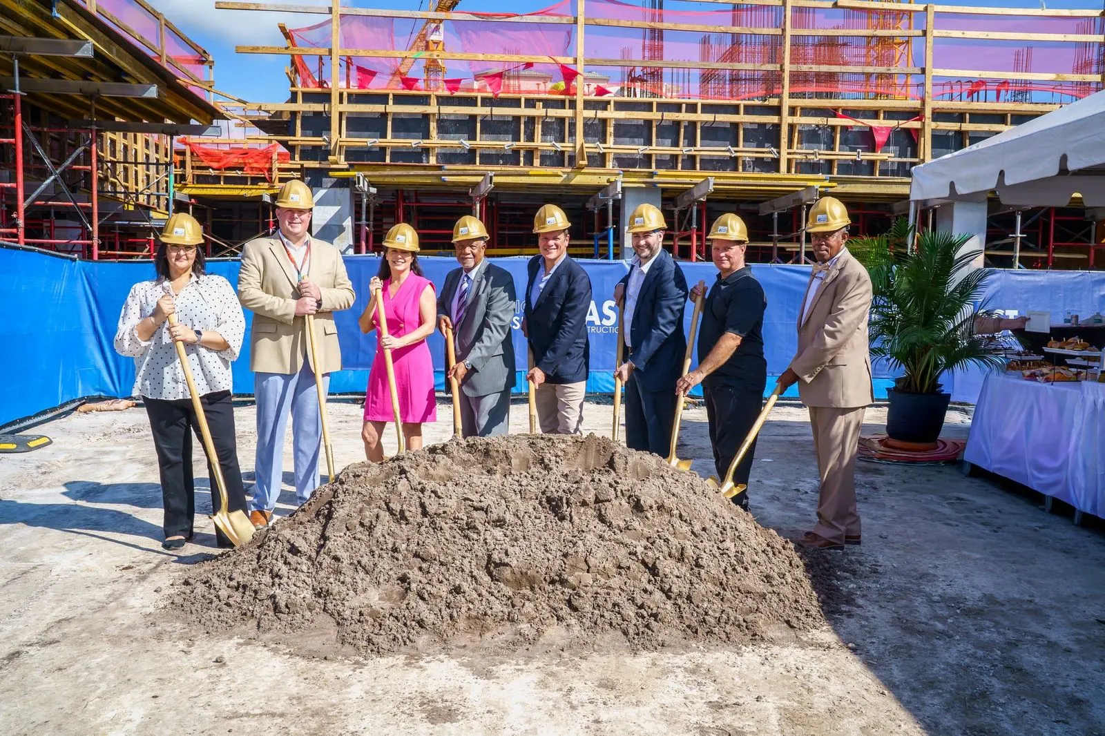 A small group of people stand with hard hats and shovels, with a building under construction in the background.