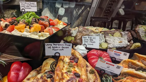 A vibrant deli display featuring colorful roasted vegetable salad, lamb pastry pies, and pork ribs with BBQ sauce, accented by fresh bell peppers.