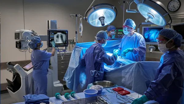 A group of people wearing blue scrubs and masks stands in an operating room. One person points to an image of lungs on a screen.