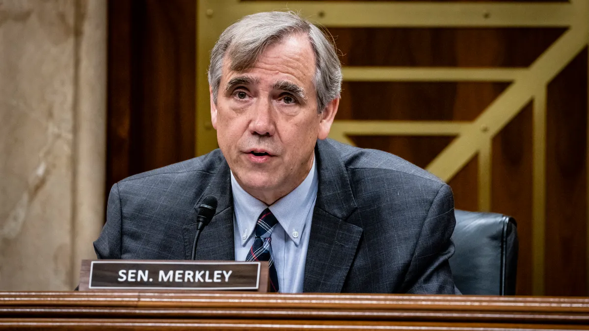 A congressman with a name placard reading "Sen. Merkley" speaks into a microphone while sitting behind a desk