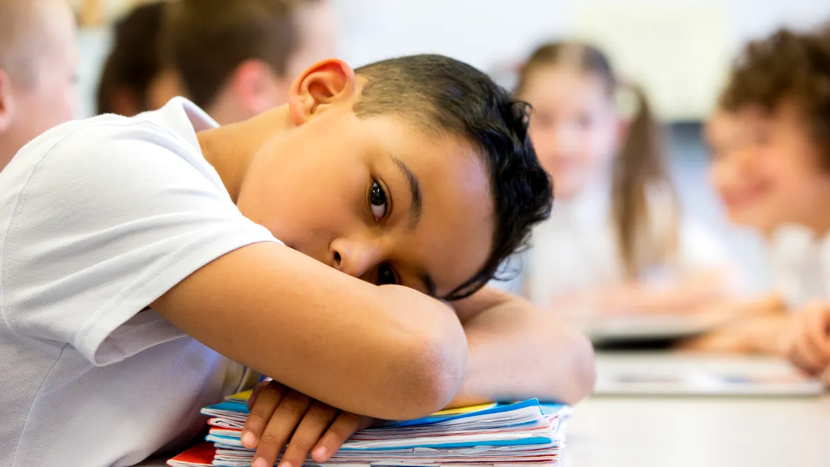 A student rests their head on their arms while seated at a desk. Under the student's arms are notebooks and the student is looking at the camera.