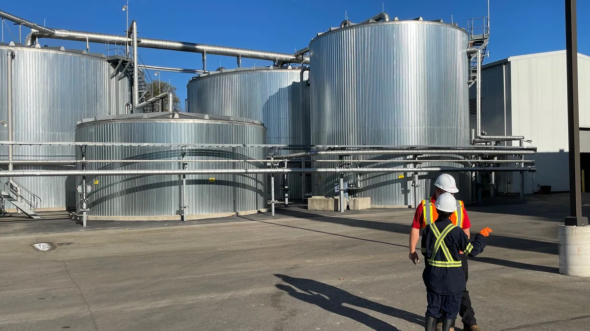 Several large cylindrical metal tanks sit outside on a concrete pad. Two people wearing protective gear talk nearby.