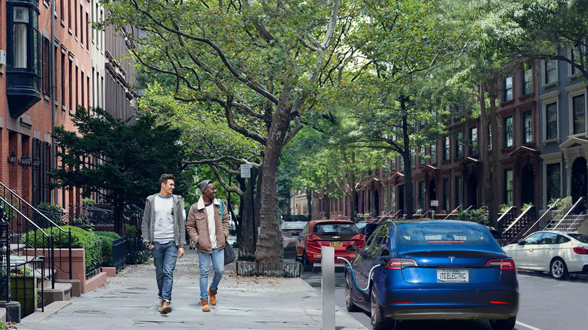 Two people walk along a street lined with classic New York brick townhouses, passing a blue car plugged into an electric vehicle charger on the sidewalk.