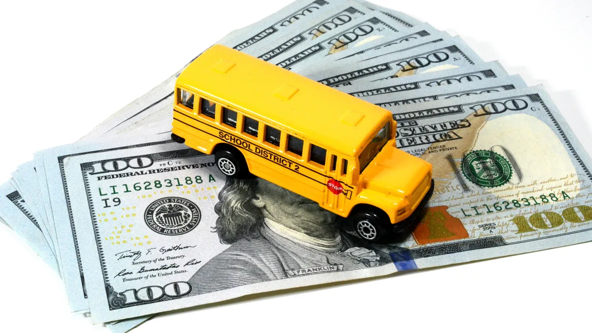 A toy yellow school bus sits on top of a fanned stack of $100 bills against a white background.