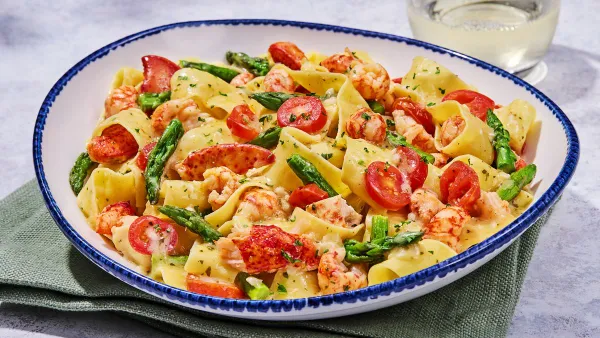 A white bowl with blue liner that contains a pasta dish with tomatoes, asapragus and lobster.