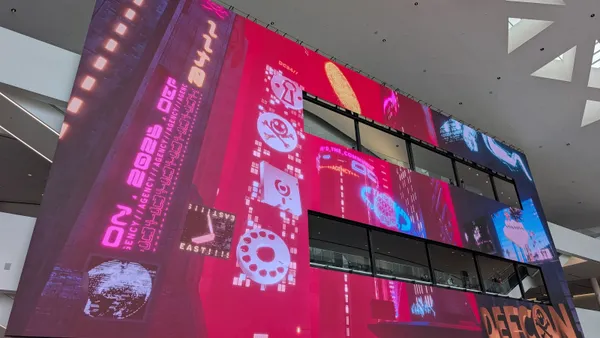 A large screen inside a convention center displays logos and other imagery related to the DEF CON cybersecurity conference
