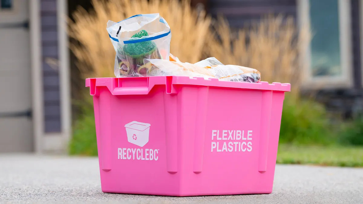 A pink recycling bin sits on the ground and  is filled with flexible plastics.