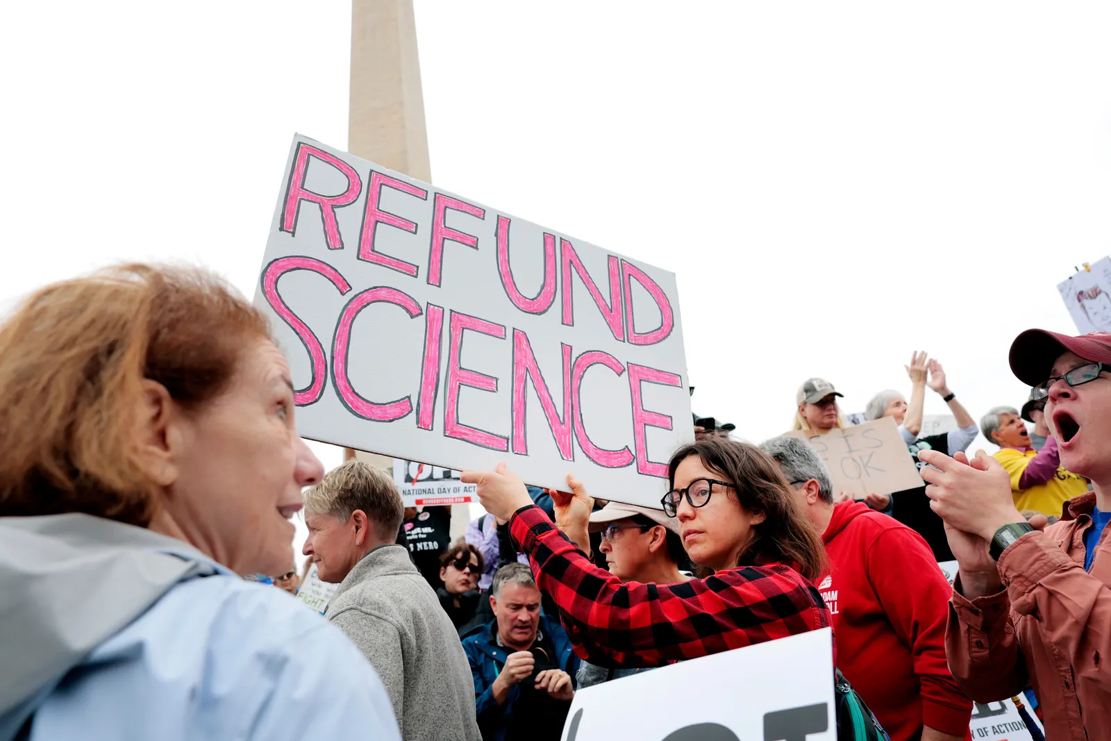 A protestor holds up a sign saying 'Refund science' in a crowd a people with the Washington Monument visible in the background.