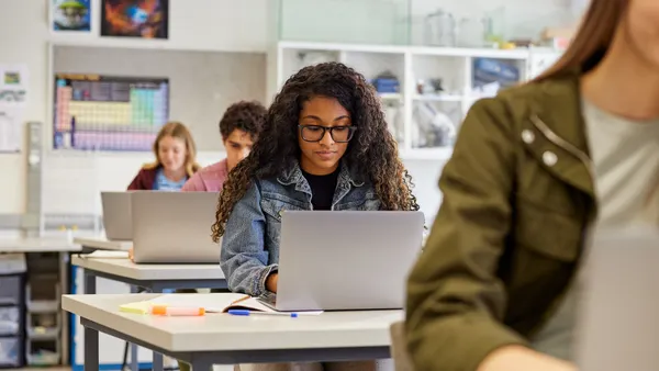 A student is pictured sitting at a desk in a classroom with an open laptop. Other students are sitting in front and behind the student.