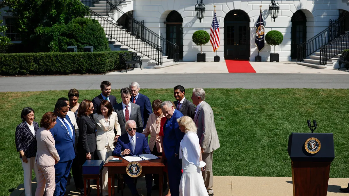 Man seated at table signs a piece of legislation, surrounded by a crowd of onlookers on a lawn outside of White House