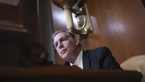 A man sits close to a microphone in a formal, wood-paneled chamber.