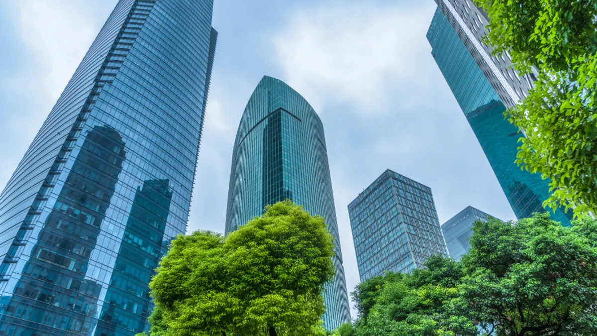 Skyscrapers with glass facades rise above green trees in a modern cityscape.