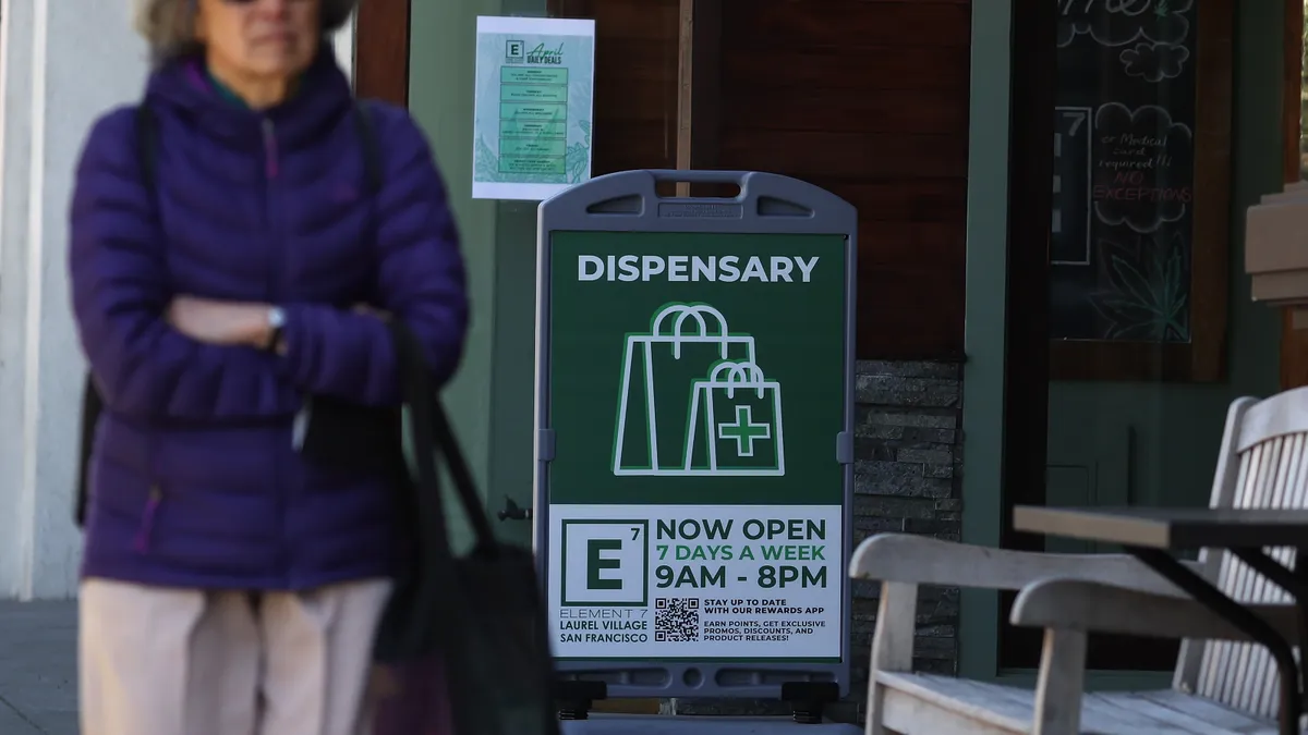 A sign in front of a dispensary shares the business's operating hours, as a person walks past.