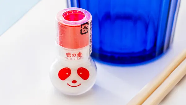A small Ajinomoto seasoning bottle with a red cap and a panda face printed on it sits on a white surface next to a blue glass, with a pair of wooden chopsticks nearby.