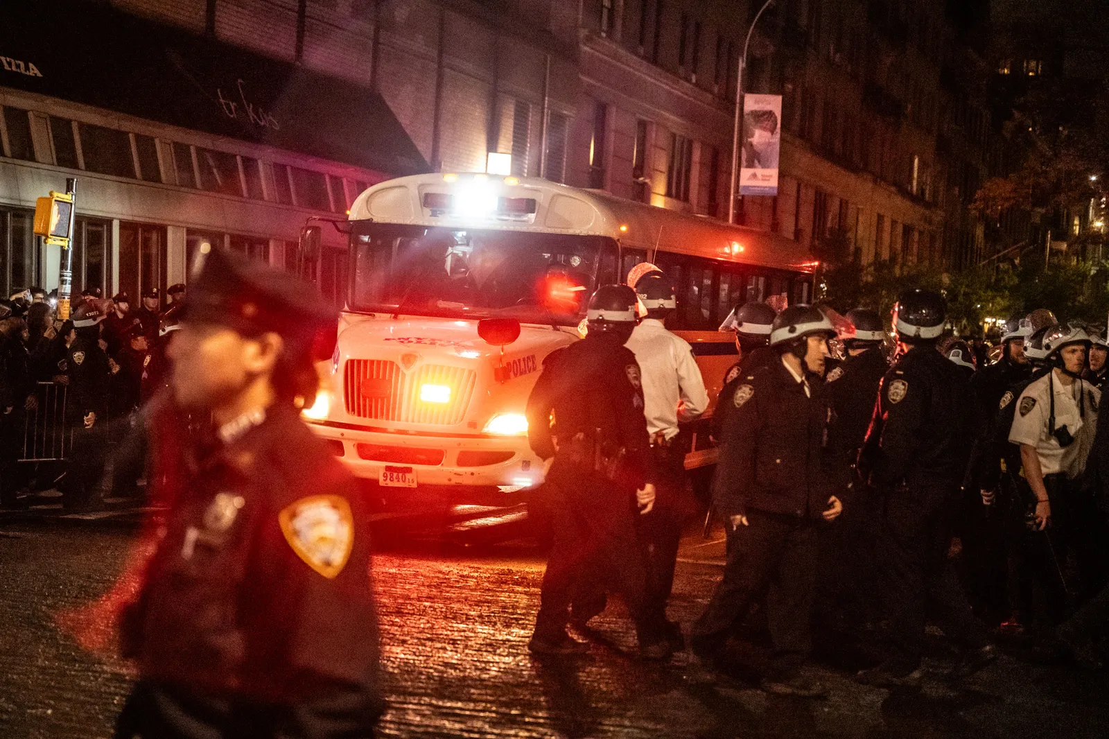 A police bus drives through a street filled with officers wearing helmets.