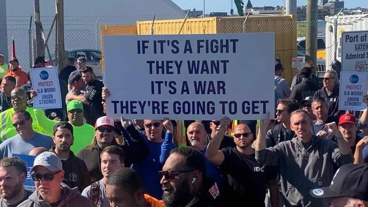 Protestors stand in a group outdoors and hold up signs. The signs read "If it's a fight they want it's a war they're going to get" and "Protect ILA work! Union strong!"