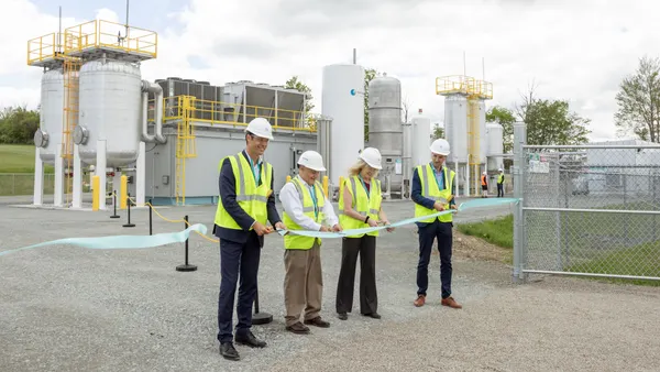 Four people wearing construction vests and hats cut the ribbon in front of a small Waga Energy industrial facility with tanks and pipes on a breezy day.