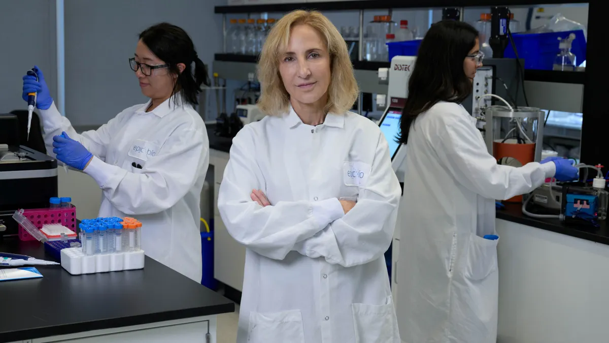a photo of Epicrispr Biotechnologies CEO Amber Salzman standing in a white lab coat facing the camera with a woman behind her to her left in a white lab coat with black glasses holding a tube and syringe, and a woman in a white labcoat behind to the right facing the right touching lab tools.
