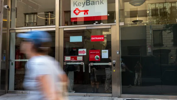 A person wearing a blue hat and light t-shirt walks past a KeyBank branch location, as the glass reflects the street scene.