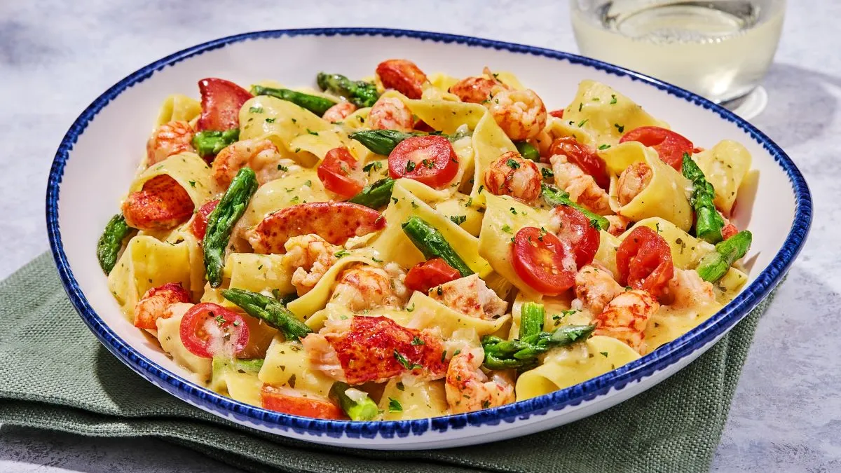 A white bowl with blue liner that contains a pasta dish with tomatoes, asapragus and lobster.