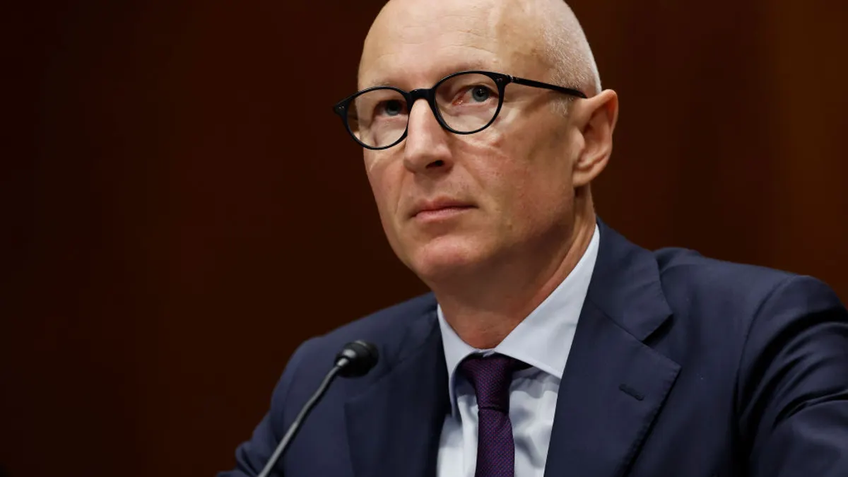A person in a suit sits at a desk with a microphone.