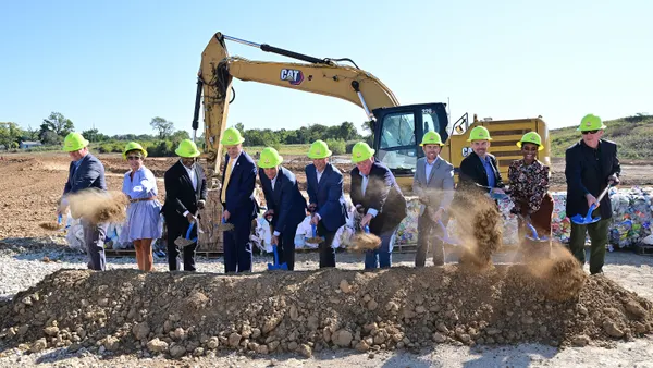 People in hard hats and business suits hold shovels as part of a groundbreaking ceremony. Heavy equipment sits behind them.