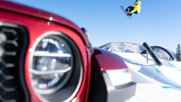 The front of a red jeep sits in the foreground while a snowboarder is mid trick in the background.