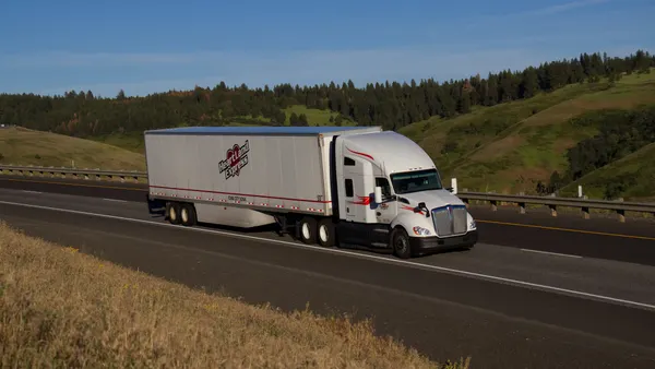 A tractor-trailer on an interstate with rolling fields and hills dotted with trees in the background.
