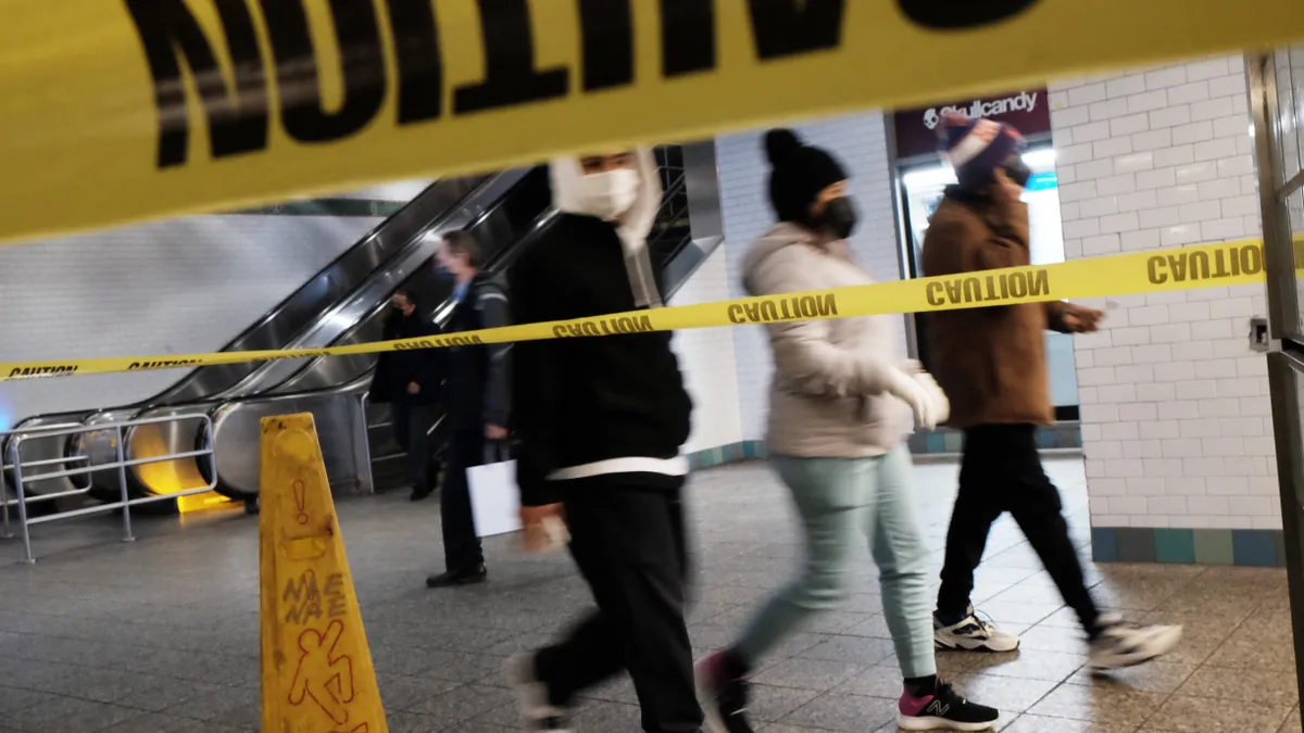 Passengers wearing masks walk through a subway stations with yellow police tape in the foreground.