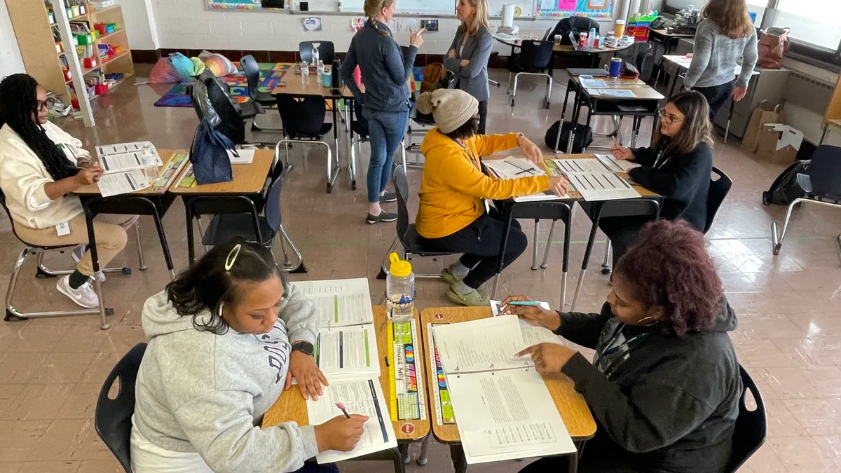 A photo of a school classroom shows adults working at individual desks with paper, pens and pencils. Several other adults are standing and talking or walking.