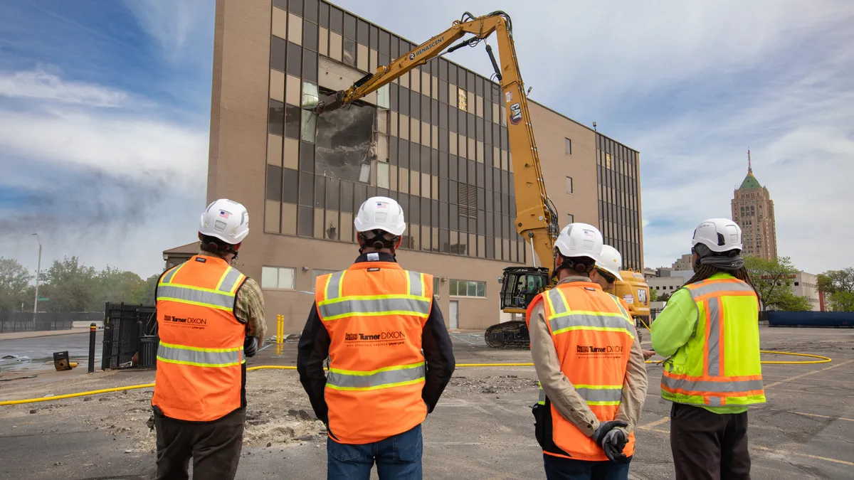 four people in orange vests that read "barton malow/turner/dixon" watch as a piece of heavy machinery punches into a building.