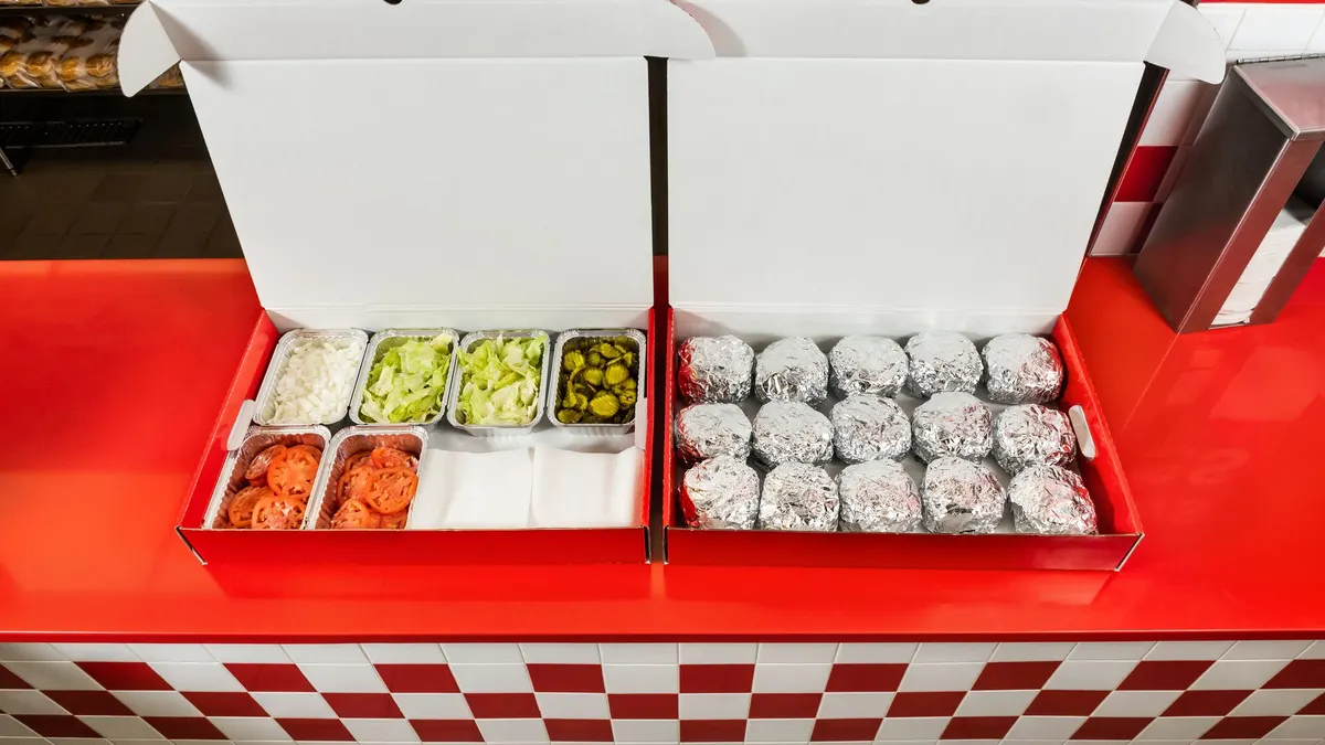 An image of two red boxes. The one on the right has wrapped burgers while the box on the left has various toppings like lettuce, pickes and tommatoes.