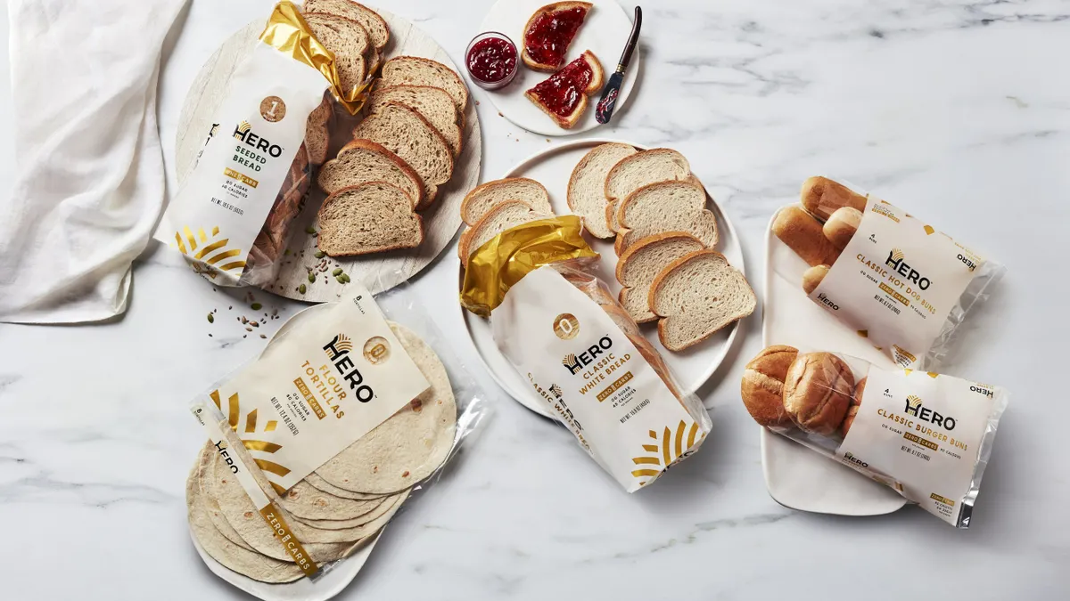 Packages of Hero Bread products, including Seeded Bread, Flour Tortillas, Classic White Bread, Classic Burger Buns and Classic Hot Dog Buns, are on plates with some products on a white marble surface.