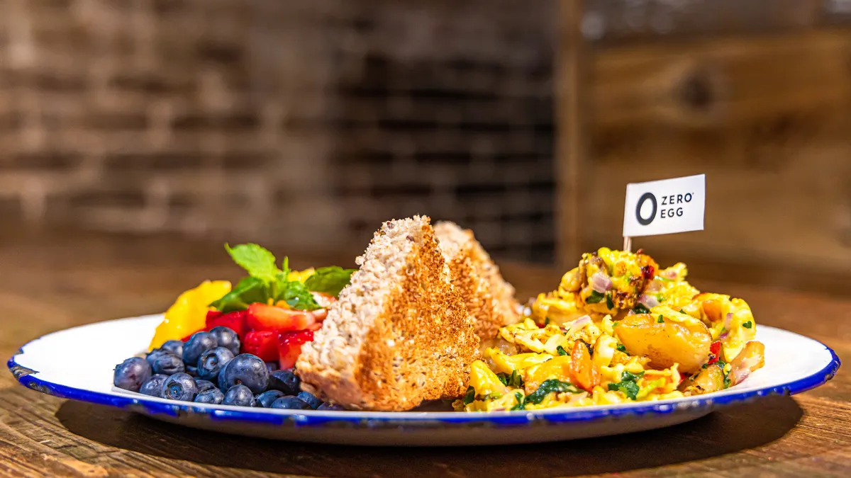 A plate containing blueberries and strawberries on the left, a toast triangle with a bite missing in the middle, and a scrambled egg with vegetables with a Zero Egg toothpick stuck in it on the right.