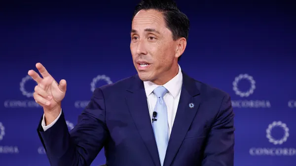 A person wearing a suit gestures with their hand while sitting in a chair in front of a blue background with Concordia labels.