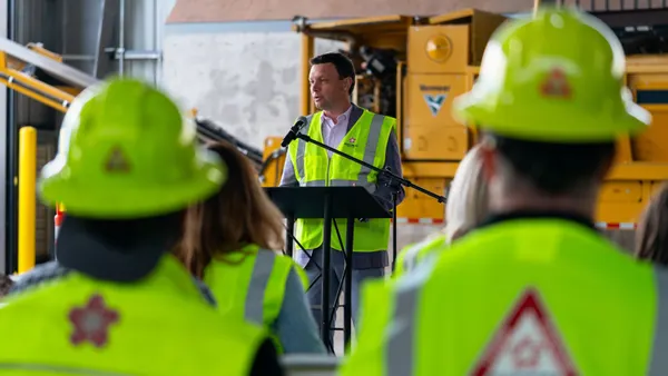 A man wearing a hi-vis vest speaks to workers wearing vests and hard hats with the Republic Services logo inside an industrial shed with construction equipment.