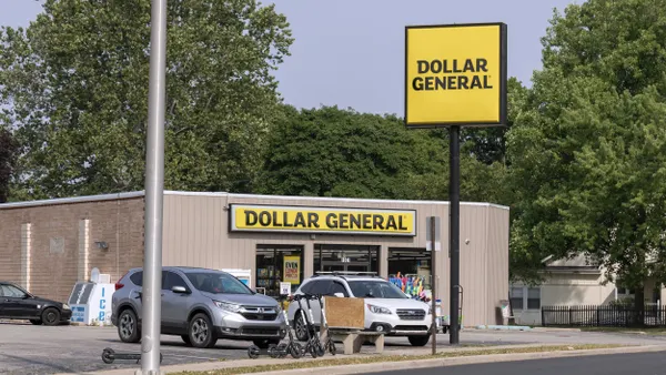 A single-story retail store with beige siding and a large yellow sign reading “Dollar General” above the entrance. Two parked cars and several electric scooters are visible in front of the building. A tall street sign with the same branding stands near the road.
