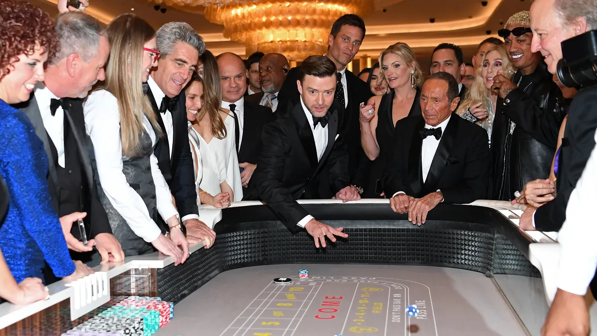 Justin Timberlake, in a suit, throws dice while surrounded by onlookers.